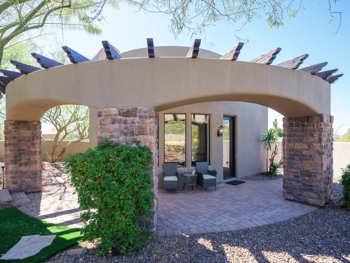 The image features an elegant exterior patio with an arched entryway supported by stone columns. Comfortable outdoor seating is arranged under dappled sunlight, surrounded by lush potted plants. The backdrop hints at serene mountain views, and the inviting atmosphere is perfect for morning coffee or evening wine.