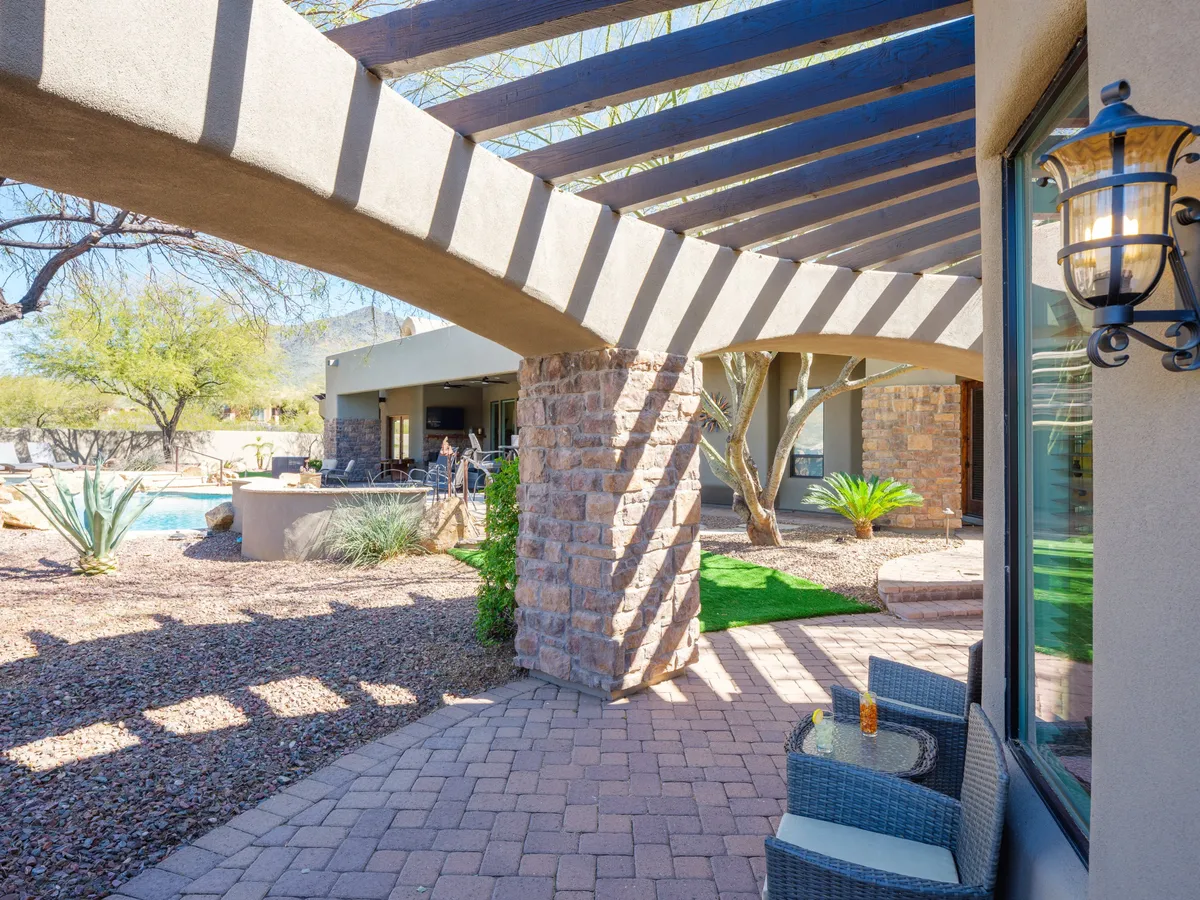 An inviting exterior setting featuring an arched pergola over a stone-columned patio, surrounded by lush landscaping with a swimming pool and outdoor seating area. The space is designed for relaxation and group gatherings, with plenty of room for socializing or hosting outdoor events.