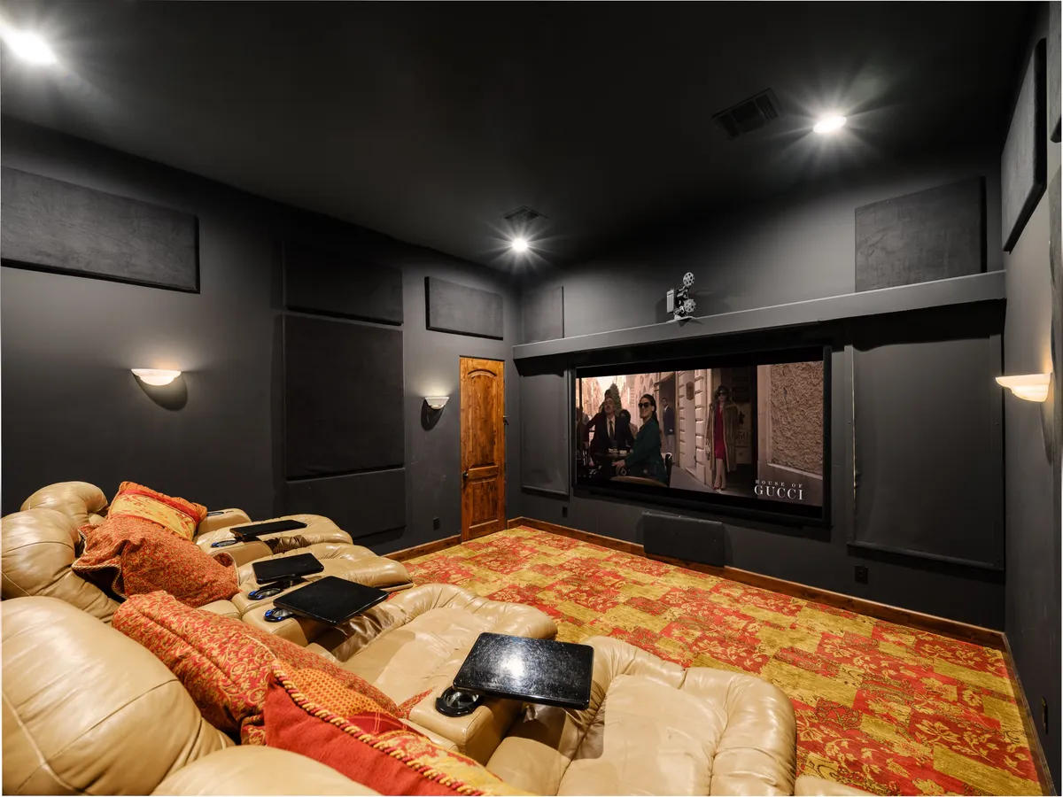 A luxurious private movie theater with plush leather seating arranged in rows facing a massive screen, decorative carpeting, and ambient lighting fixtures that set a cinematic mood. The room is purpose-built for immersive movie nights and intimate gatherings.