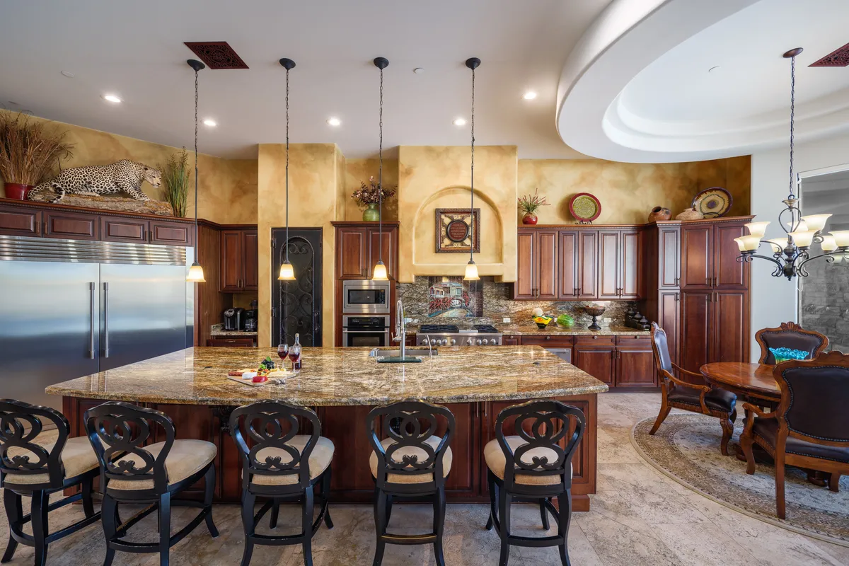A luxurious kitchen scene with a granite island at its center, stainless steel appliances, wood cabinetry, and chandeliers. Decorative plants and expansive counter space invite cooking, entertaining, and family gatherings.