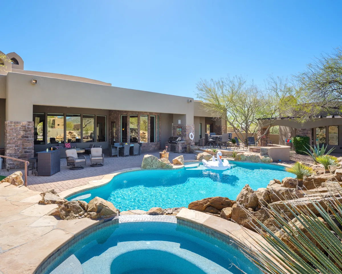 The image showcases the home's sparkling swimming pool surrounded by elegant outdoor seating, lush greenery, and distinctive desert rock formations, all set against the stunning backdrop of the Cave Creek mountains. The scene evokes relaxation and luxury, with inviting spots for sunbathing, outdoor dining, and unwinding as the sun sets over the landscape.