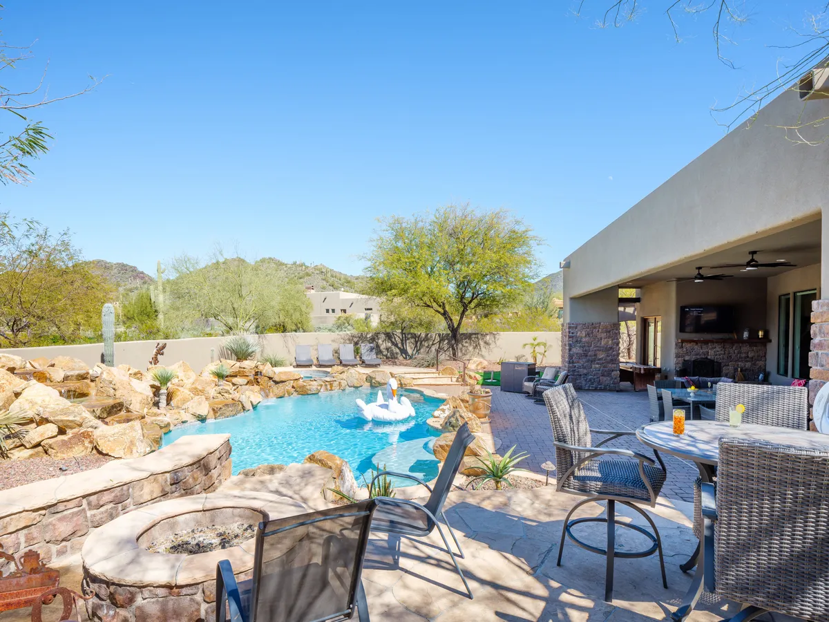 A tranquil, resort-style backyard features a sparkling swimming pool, comfortable outdoor seating, unique rock formations, and lush landscaping. The patio area is inviting, perfect for swimming, relaxing, and family gatherings under the sun or stars.