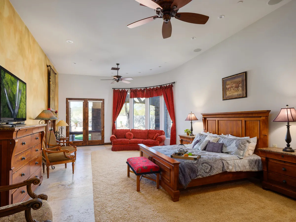 A spacious bedroom featuring a large bed with decorative bedding, a red sofa for additional seating, and classic wooden furniture. The room is warmly lit and designed for comfort, offering a welcoming space to relax and unwind. A television is positioned for easy viewing from the bed or sofa, and the overall decor exudes a cozy, restful mood.