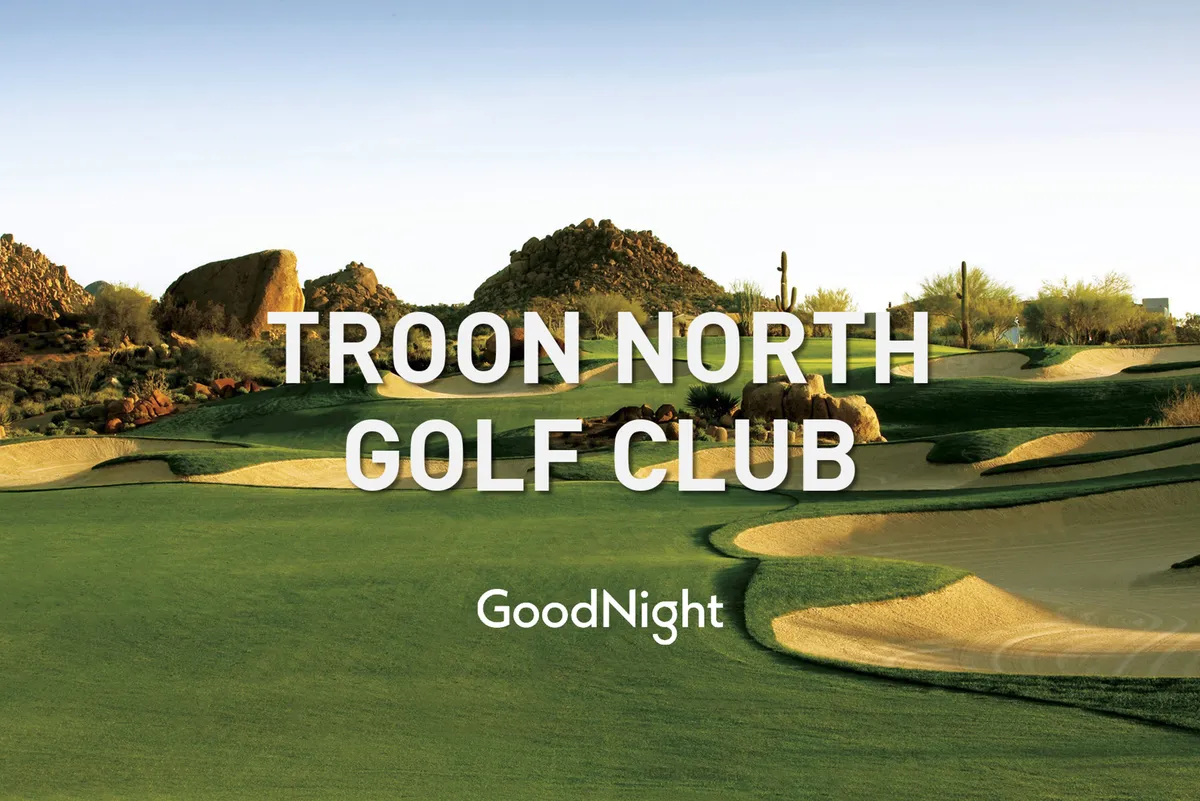 A lush golf course unfolds in the foreground, framed by dramatic rock formations and native desert greenery. The landscape extends toward distant mountains under a vibrant sky, evoking the region’s signature blend of golf and natural beauty.