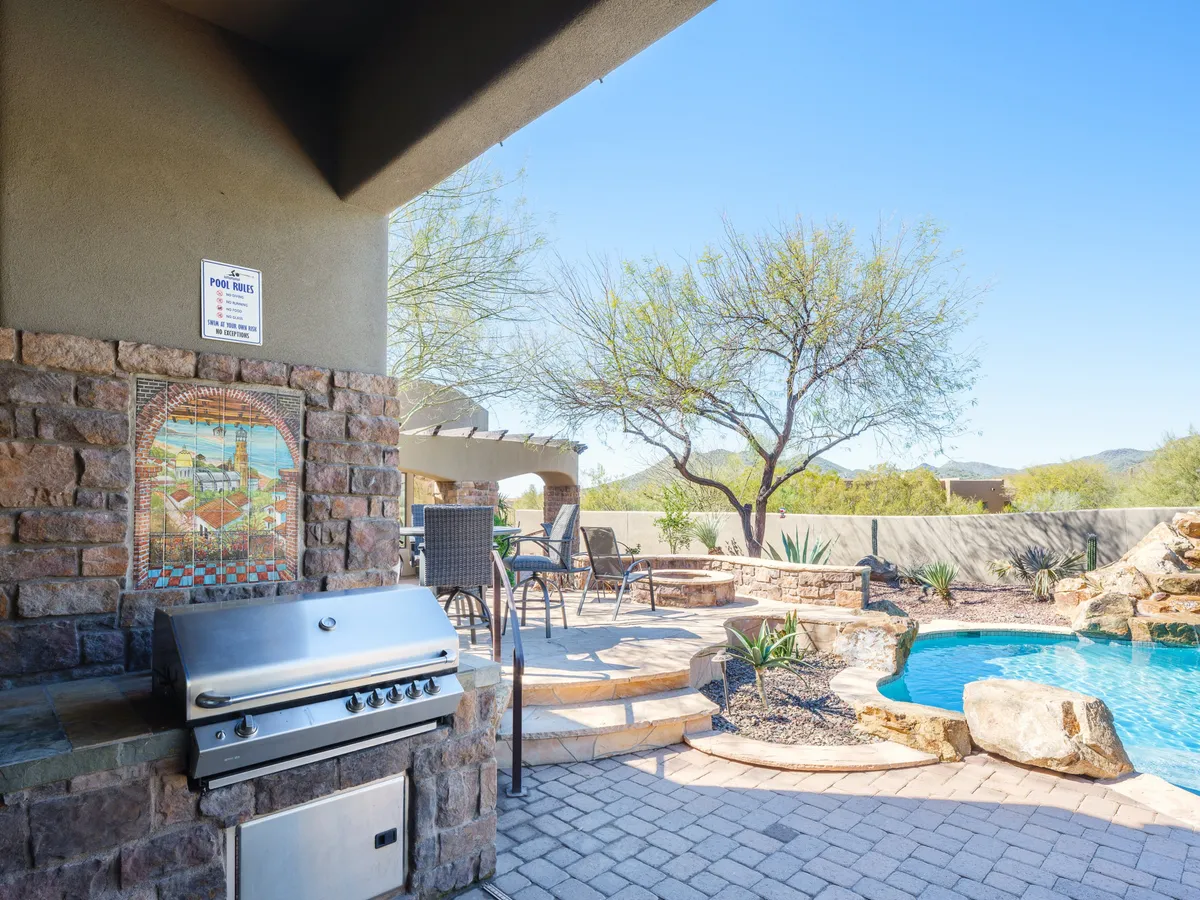 A festive outdoor patio scene featuring a barbecue grill, a sparkling pool nearby, comfortable patio seating, and colorful mosaic artwork. The setting is perfect for lively gatherings, pool parties, and al fresco meals under the sunset.